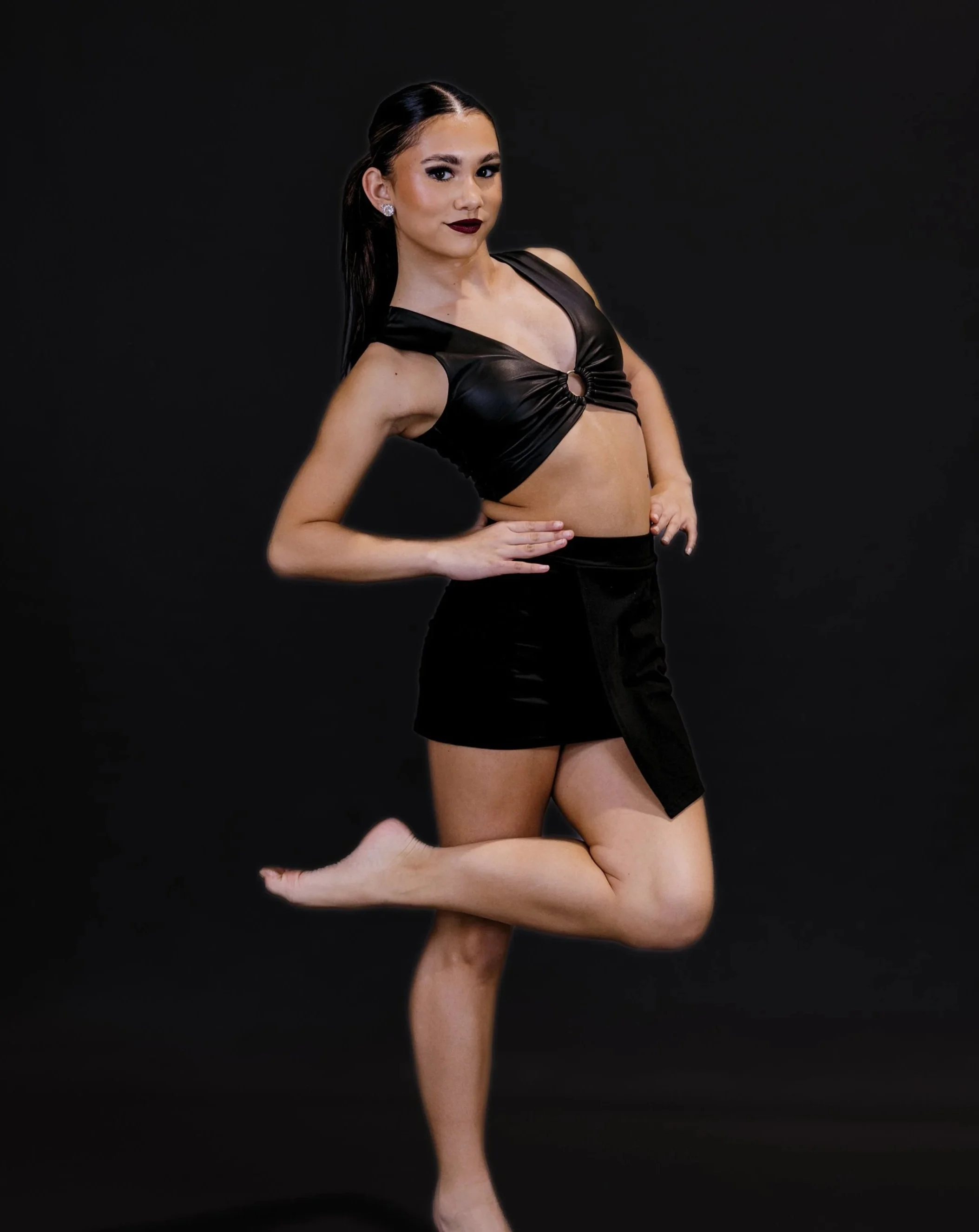 A woman with long dark hair, wearing dark makeup and a black leather crop top, posing with one knee bent against a plain black background.
