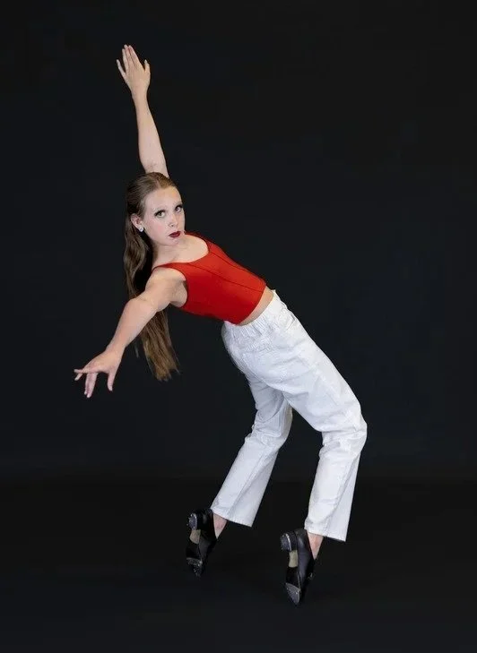 A young woman in a red top and white pants performs a dance pose against a black background.