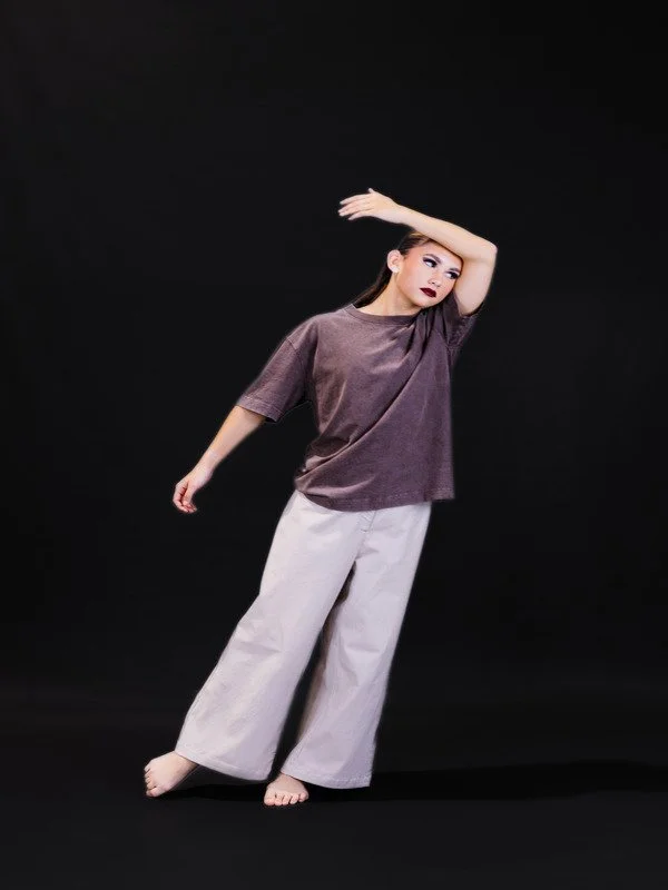 A young woman with long dark hair against a black backdrop, wearing a loose purple t-shirt and wide white pants, striking a pose with one arm raised above her head and the other relaxed by her side.