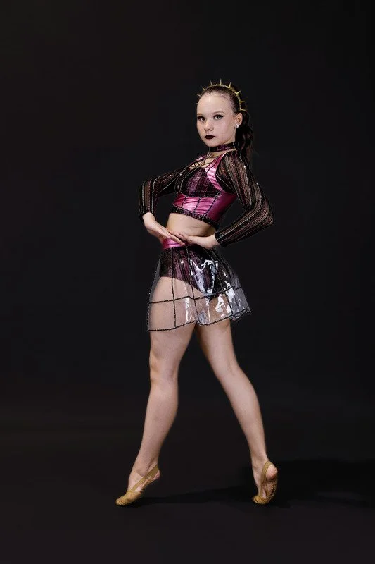 A young girl with a confident pose wears a fashionable outfit, including a sheer black and pink top, a clear skirt, and gold heels, standing against a black background.