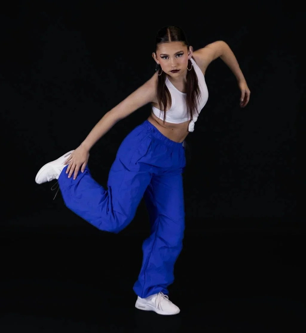 Young woman in a dance pose, wearing a white crop top, blue baggy pants, white sneakers, and hoop earrings against a black background.