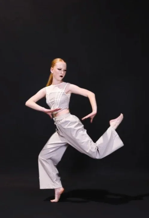 A dancer performing in a studio with a black background, wearing a white sleeveless top and light-colored loose pants, striking a dynamic pose with one foot on the ground and the other lifted, arms positioned gracefully.
