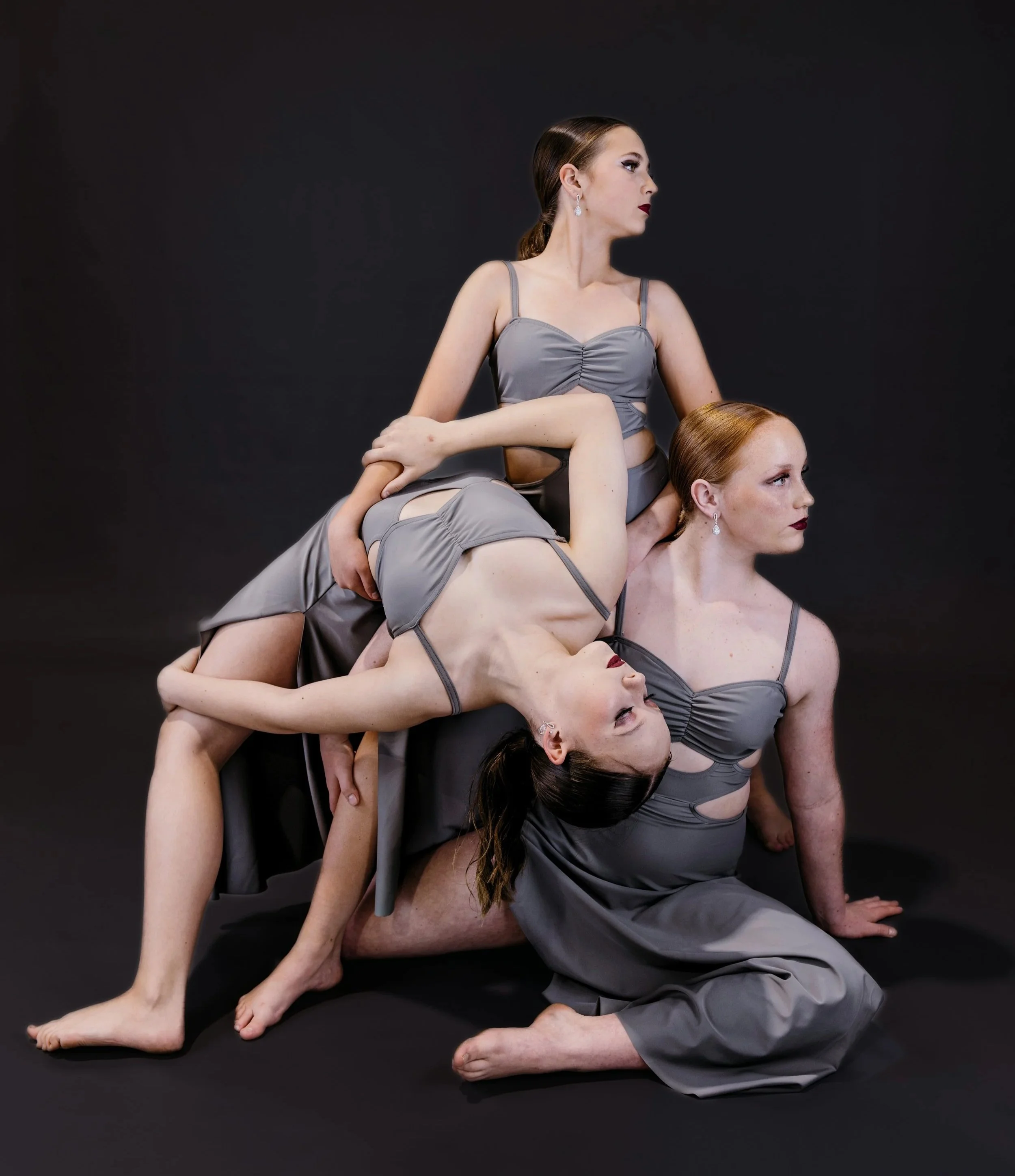 Three women in gray dresses are stacked in a yoga pose against a black background.