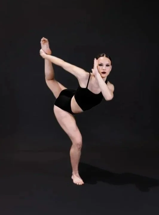 A woman performing a balancing yoga pose against a black background, standing on one leg while holding the other behind her with one hand, and placing the other hand near her face.
