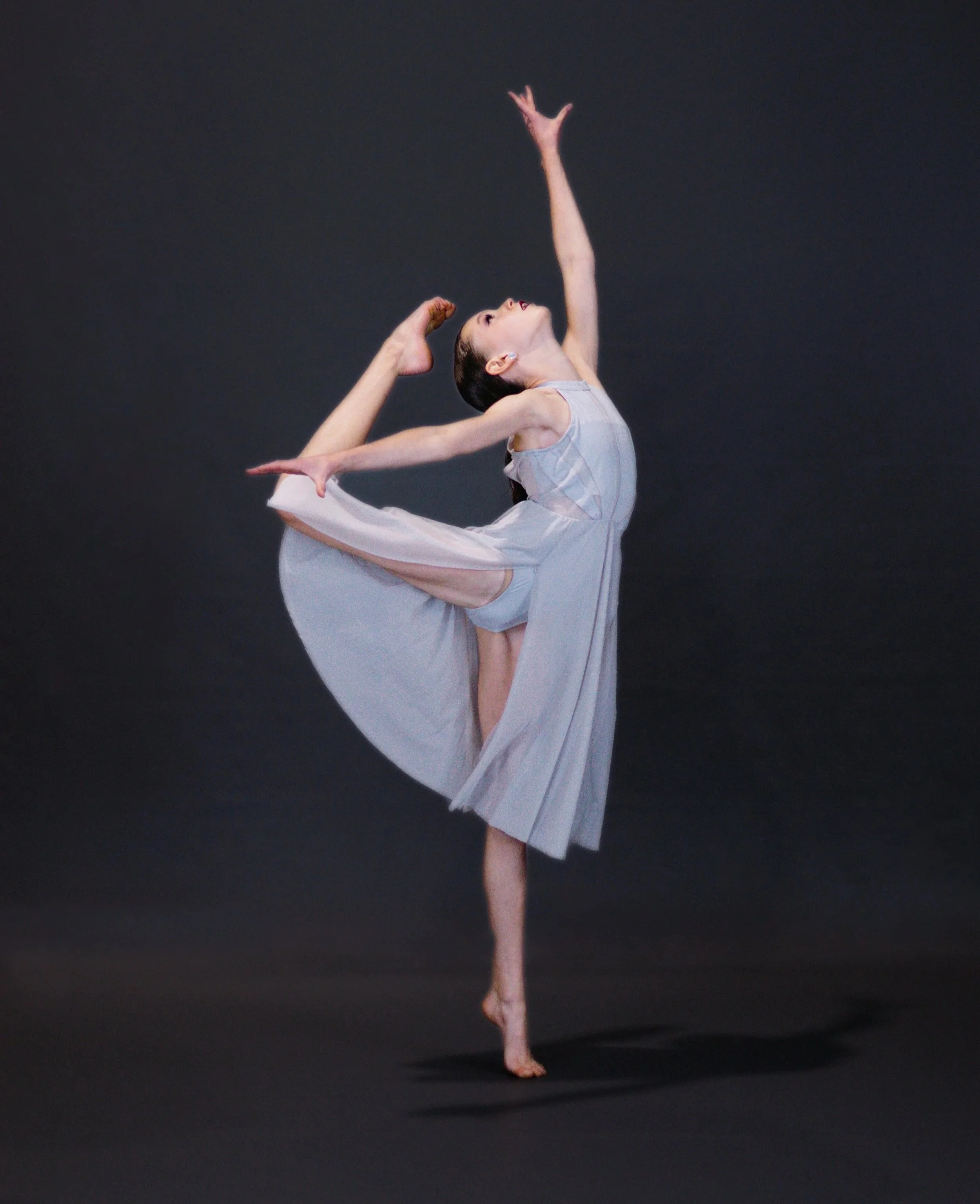 A ballet dancer in a pose with one leg raised high and arms extended, wearing a light gray dress against a dark background.