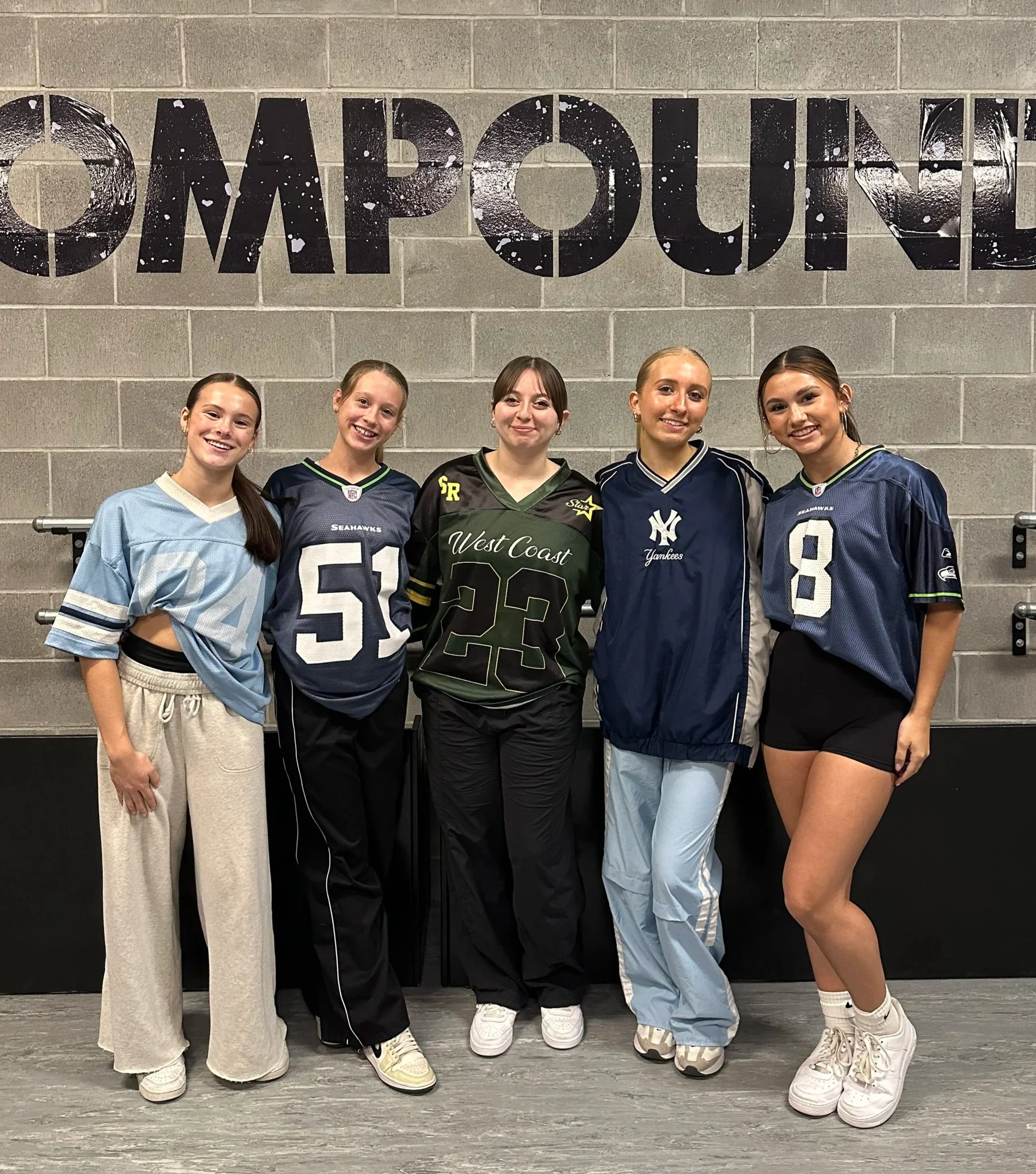 Five young women standing in front of a gray cinder block wall with the word 'DAPOUI' painted on it. They are all smiling and wearing sporty outfits, with sneakers and casual pants or shorts.