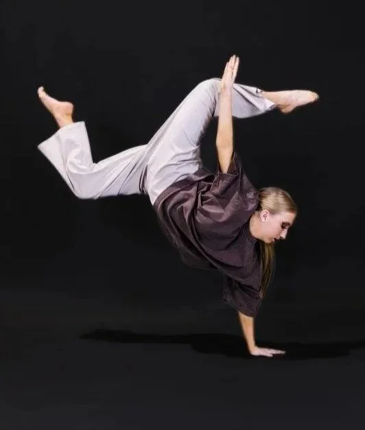 A woman in gray sweatpants and a dark top performing a handstand where her legs are bent and raised, with one hand on the ground and the other arm reaching upwards, against a black background.