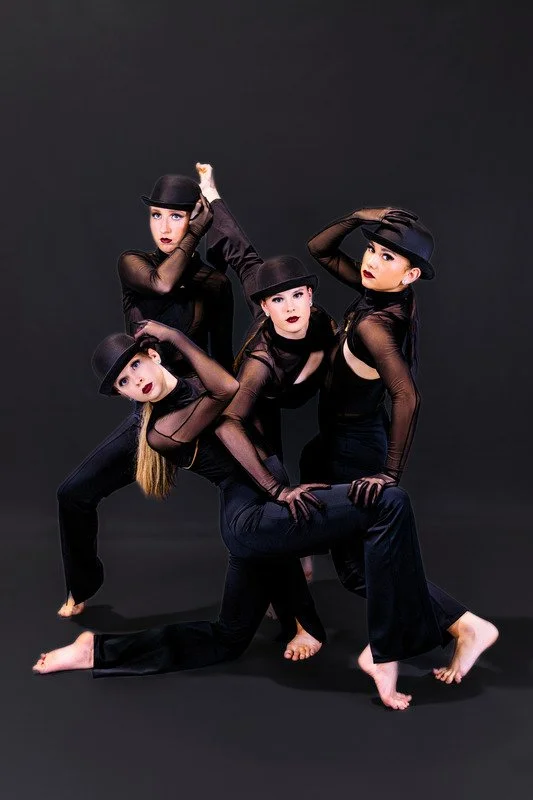 Four women in black dance outfits and hats posing against a black background.