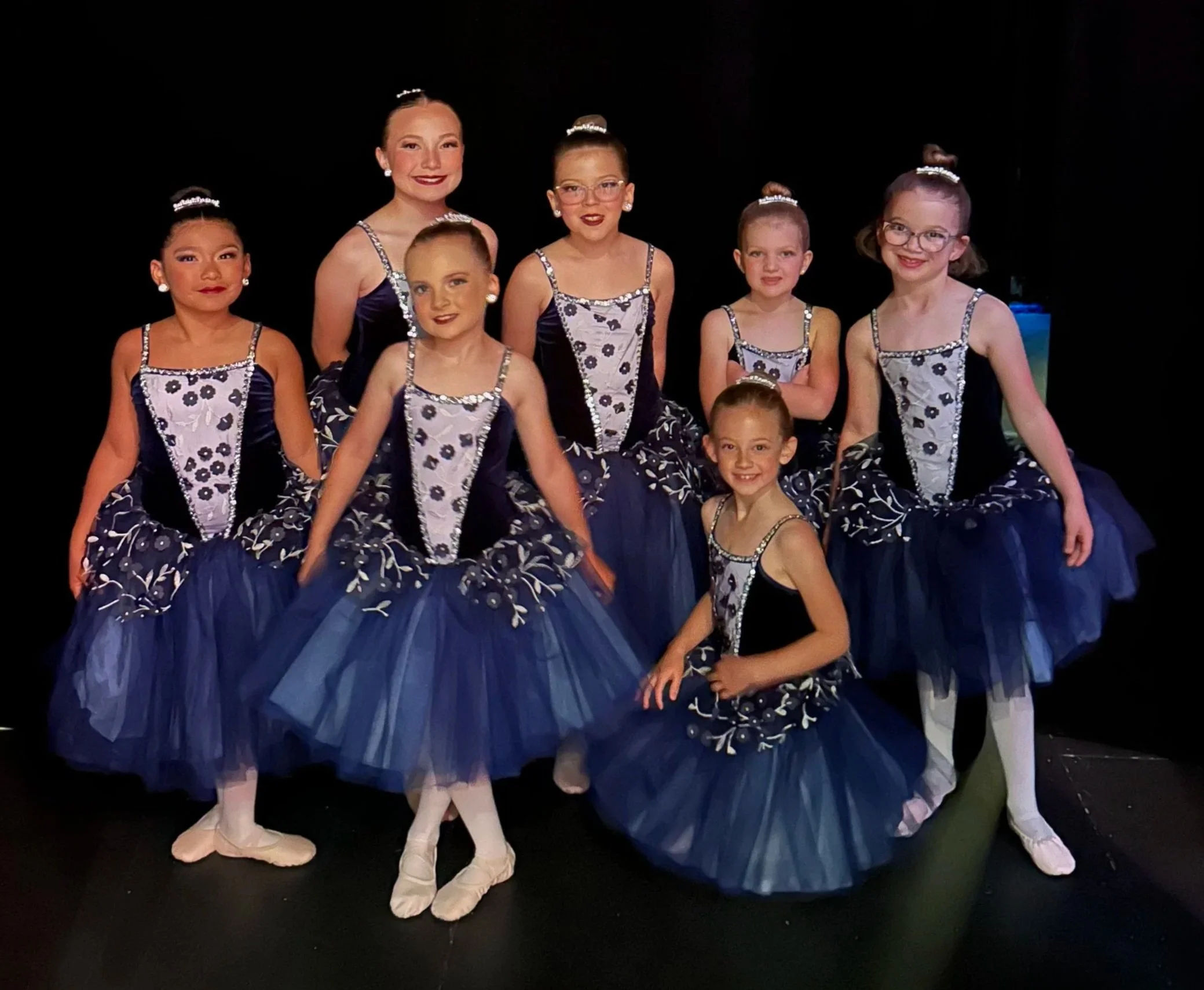 Group of young ballet dancers dressed in matching navy and white tutus with floral patterns, wearing ballet slippers and pearl earrings, standing together backstage with a dark background.