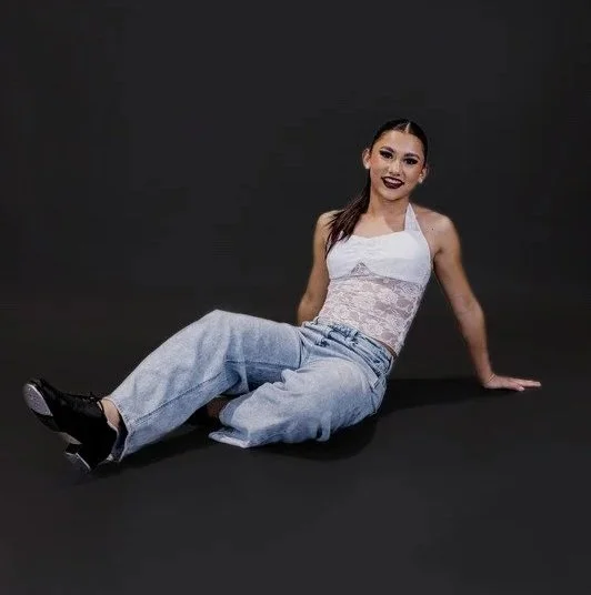 A young woman with dark lipstick and makeup sitting on a black floor against a dark background, wearing a white lace top and light blue wide-leg jeans.