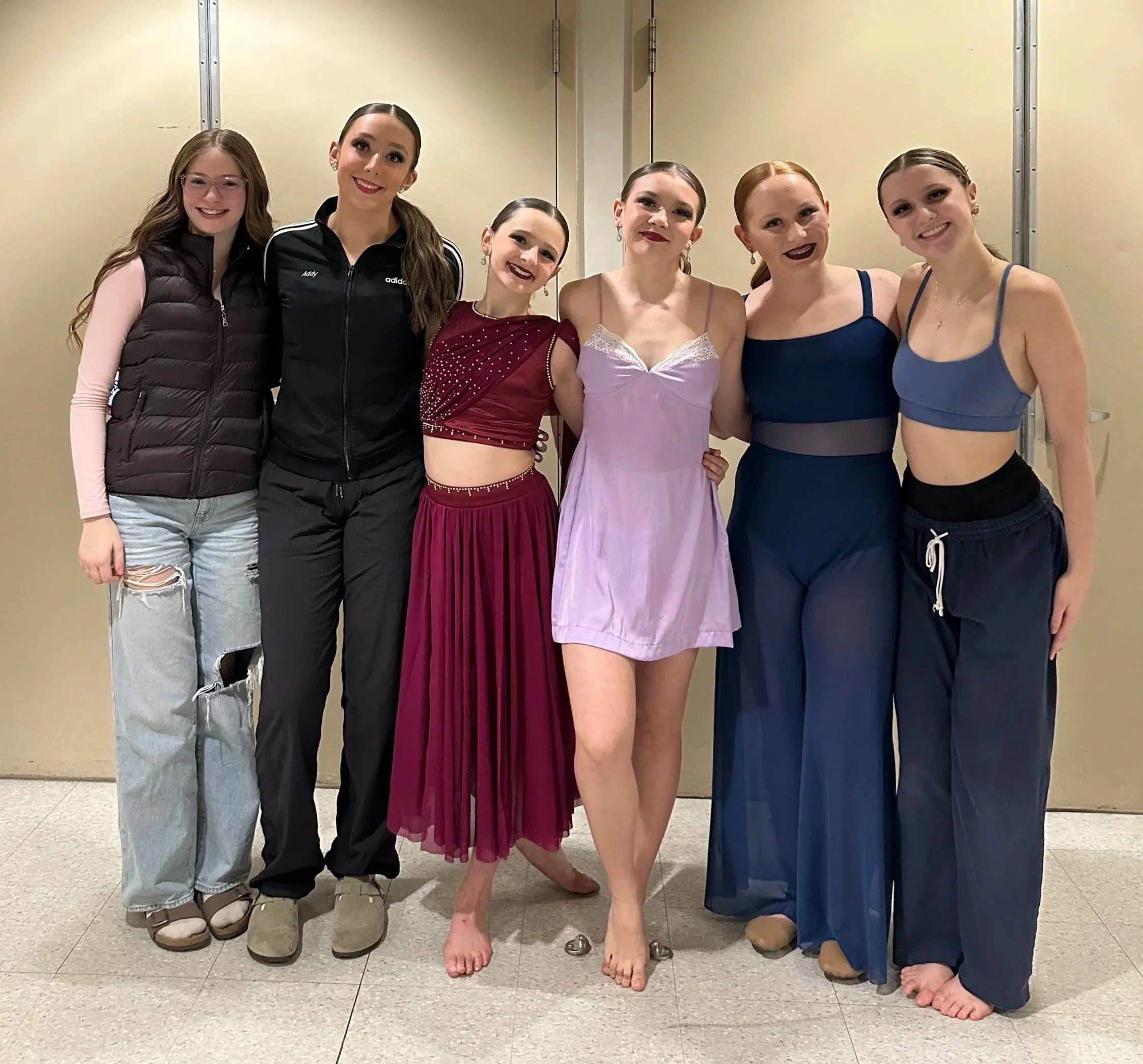 Group of six young women standing close together, smiling for the camera in an indoor setting with plain beige walls; one woman is dressed in an elegant ballet costume, and the others are casually dressed, with one wearing a jacket labeled 'Adidas'.
