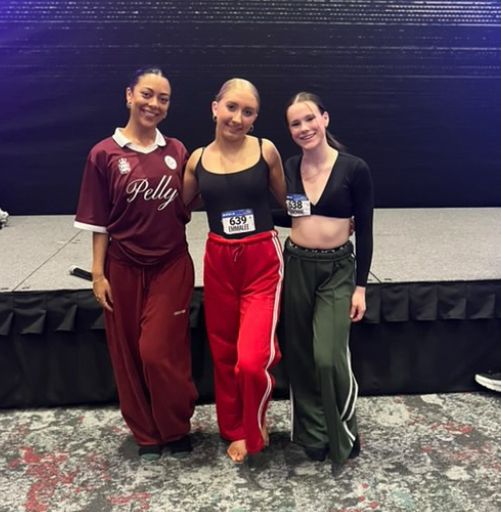 Three young women standing together on a stage, smiling, dressed in athletic clothing and wearing race bibs, at an indoor event.