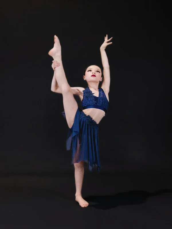 A young girl in a blue dance costume performing a high leg stretch with one arm raised and the other holding her raised leg against her head, against a black background.