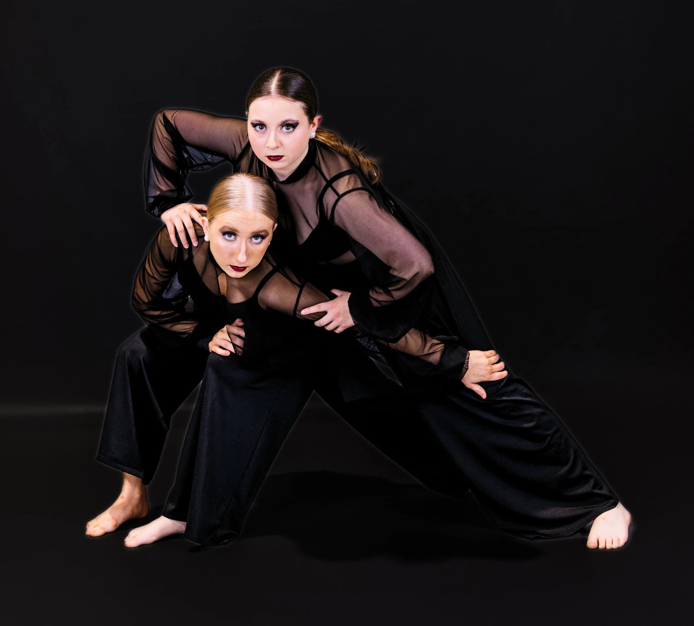 Two women performing a dance pose against a black background, wearing black sheer dresses, with one woman crouching and the other leaning over her.