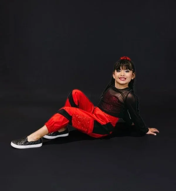 Young girl sitting on a black background, wearing a black and red outfit with sparkly shoes, smiling at the camera.