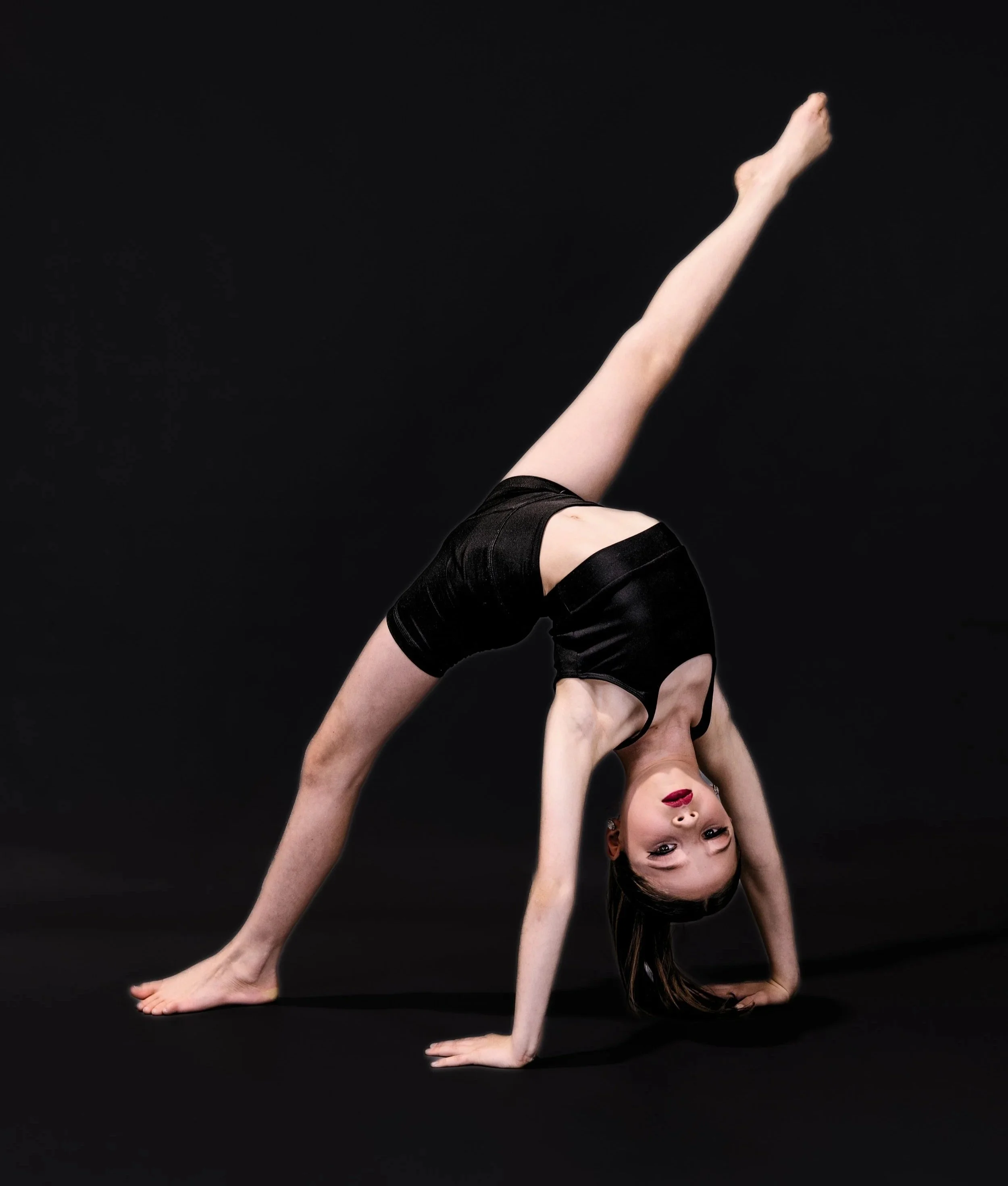 A woman performing a backbend yoga pose on a black background, wearing black shorts and a black top, with long brown hair and red lipstick.