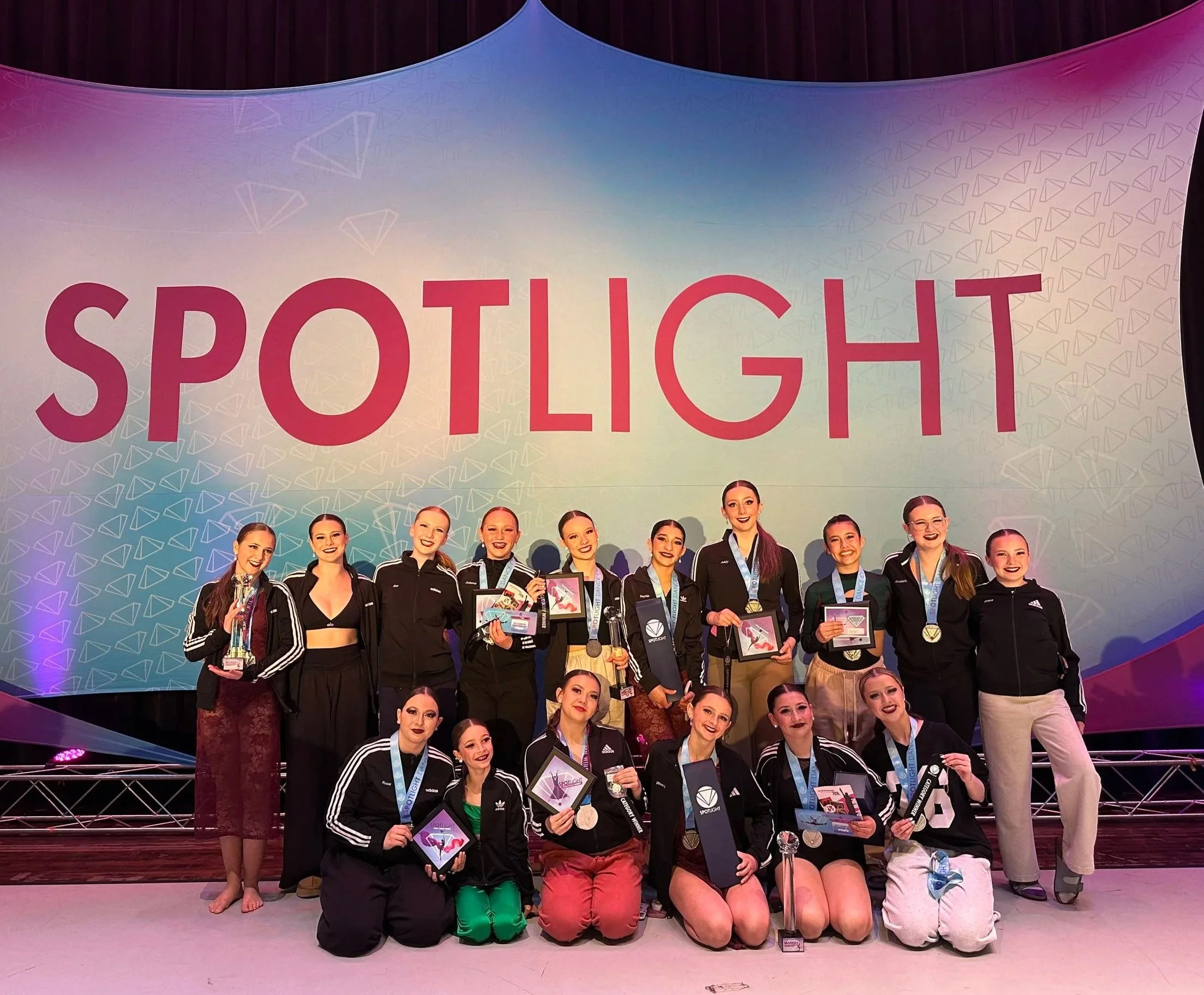 A group of female athletes standing on a stage after a competition. They are dressed in black athletic jackets with white stripes, and some are holding medals, certificates, and awards. The background features a large banner with the word 'SPOTLIGHT' in bold pink letters and a pattern of diamond shapes.