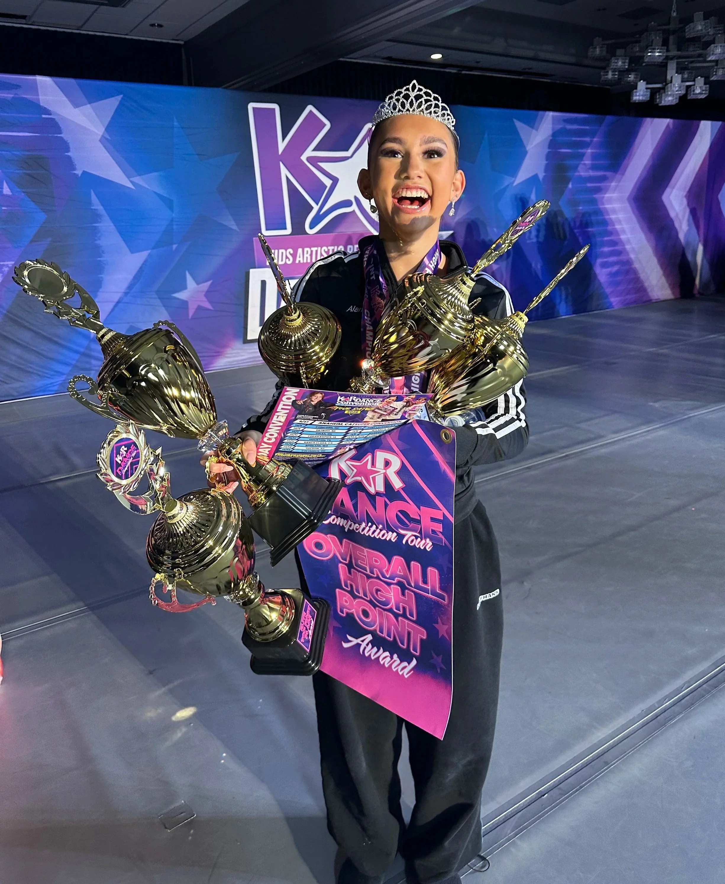 Young girl wearing a tiara and medals, holding trophies and a large award banner, at a dance competition. She is smiling with her mouth open, in front of a stage backdrop with the logo 'KAR' and star designs.