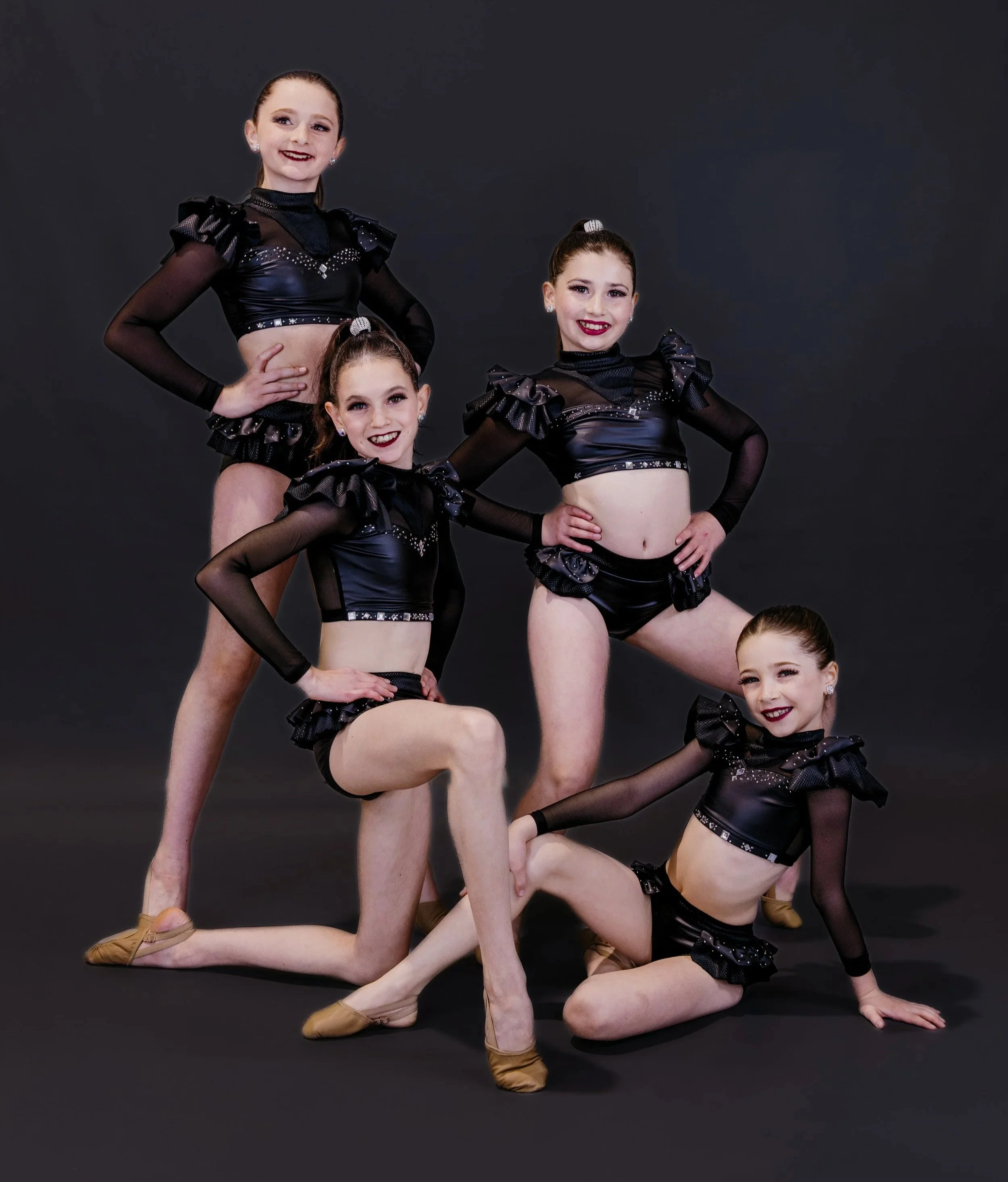 Four young girls in black dance costumes with ruffled sleeves and shiny embellishments, posing on a black background, one girl kneeling and the others standing with hands on hips.