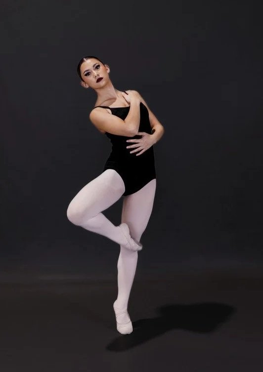 A female ballet dancer in a black leotard and white tights standing on one leg with the other leg bent and foot resting on her knee, against a dark background.
