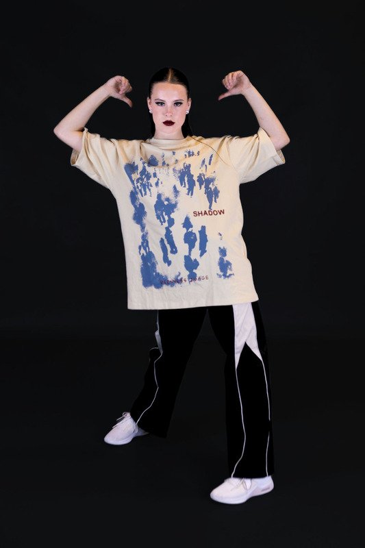 Person standing against a black background, wearing a beige oversized T-shirt with blue and red text and graphics, black pants with white stripes, and white sneakers, flexing their arms.