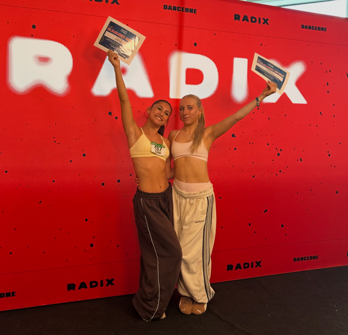 Two young women in athletic wear standing in front of a bright red backdrop with the words 'RADIX DANCEONE' on it, smiling, and holding certificates above their heads.