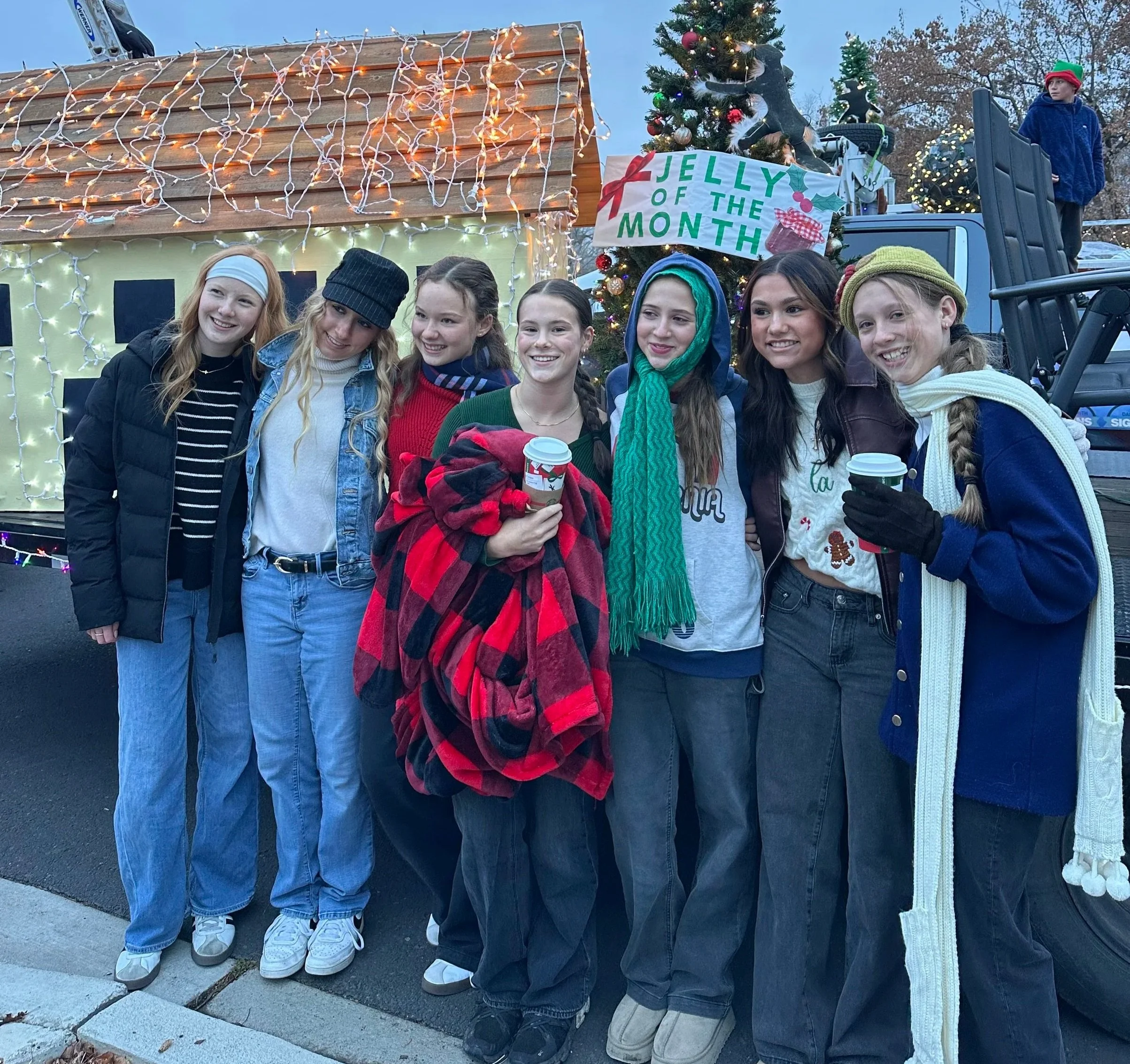 Group of young girls smiling in front of a decorated float with Christmas lights and a Christmas tree, with a banner that reads 'Jelly of the Month'.