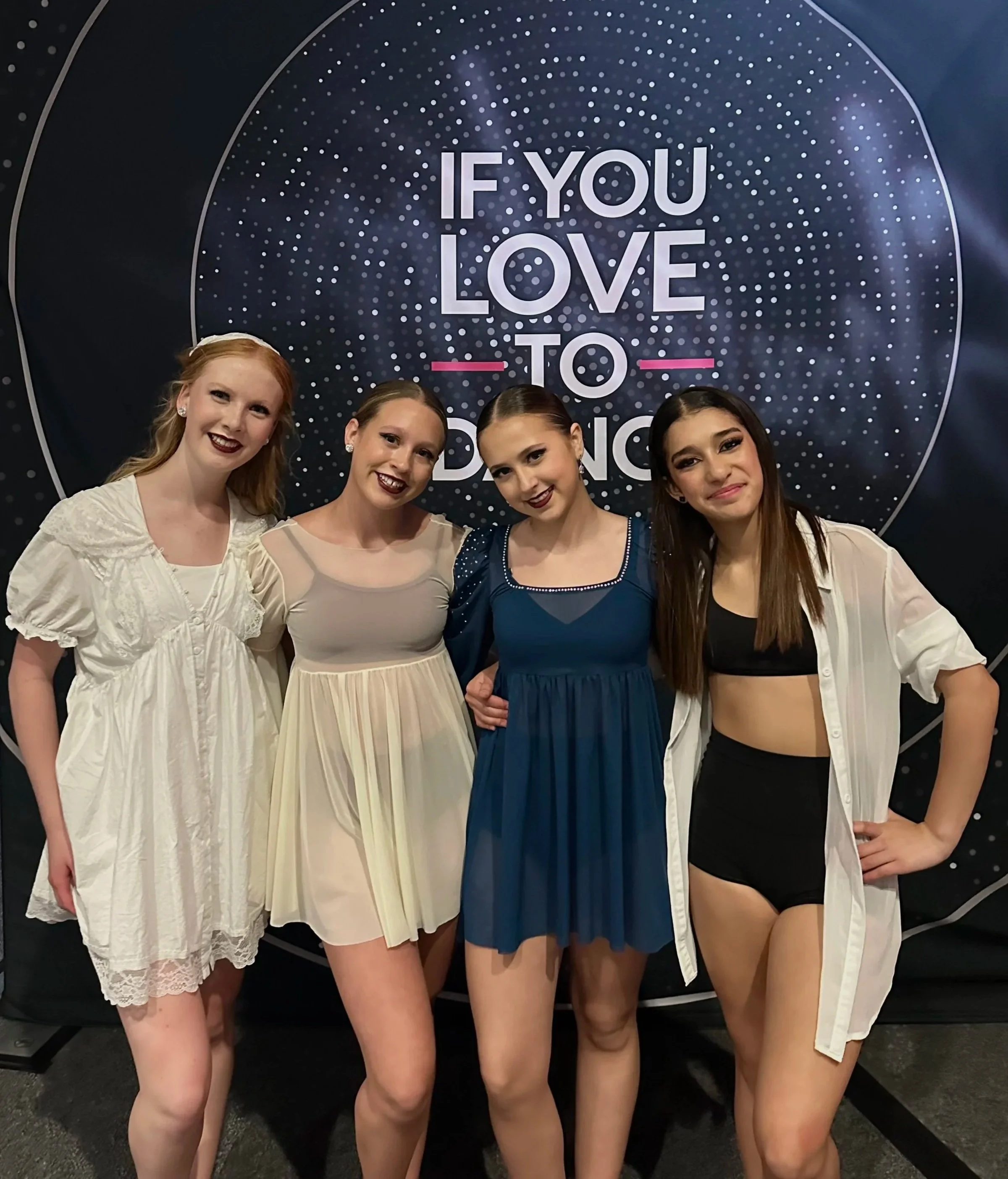 Four young women dressed in dance costumes standing in front of a backdrop with the words "If You Love To Dance" surrounded by a dotted circular design.