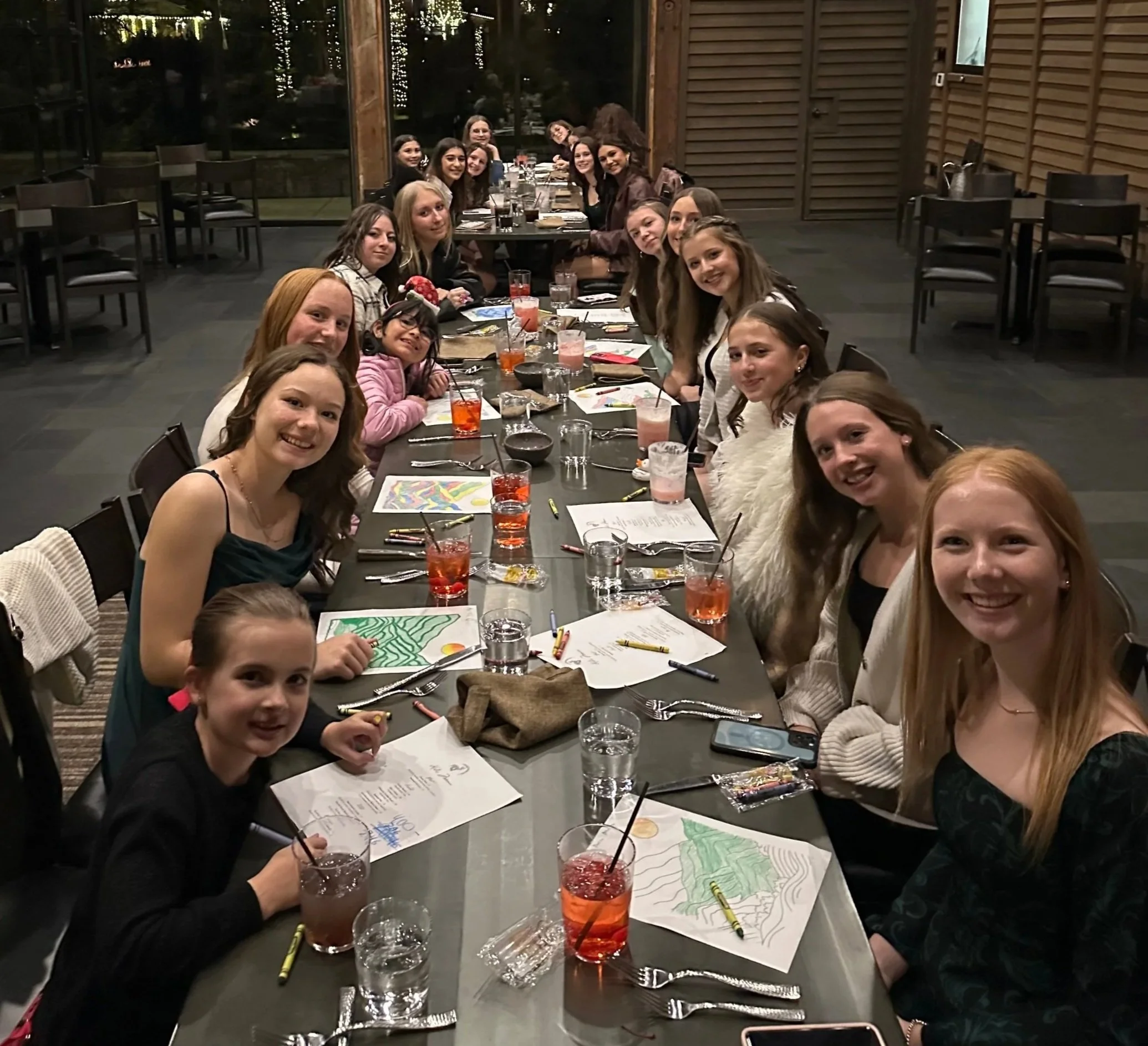 A group of young women and girls sitting around a long table in a dimly lit restaurant, smiling at the camera. The table is set with drinks, papers, and art supplies, suggesting they are participating in an art activity or celebration.