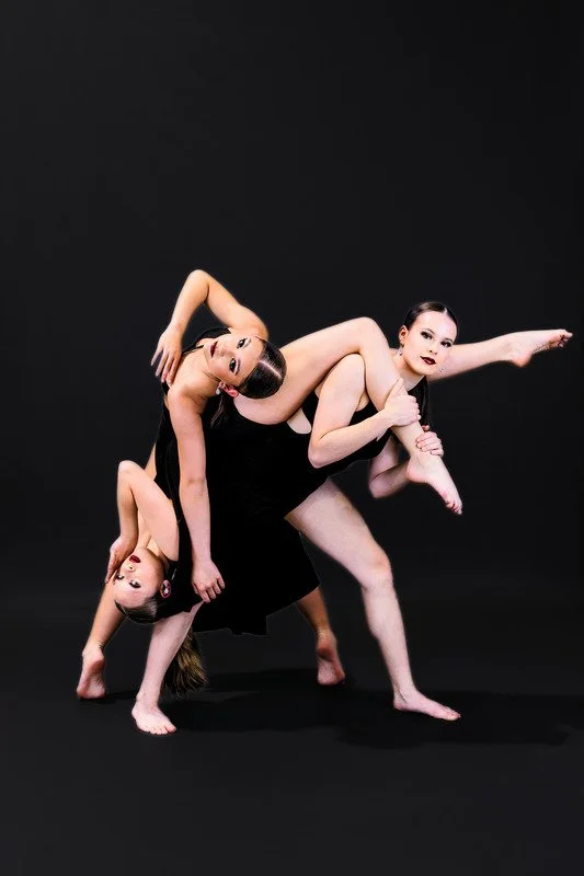 Three women in black dresses performing a contemporary dance or pose against a black background, with one woman holding a child and another woman balancing on her.