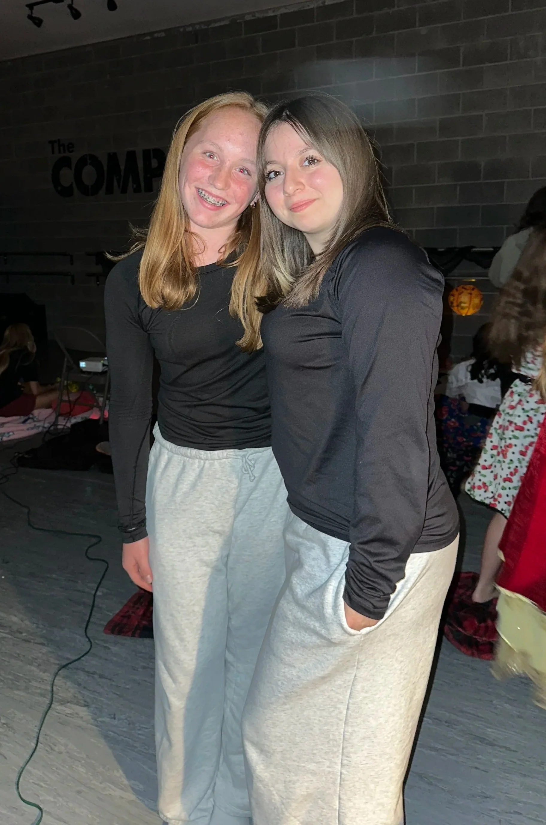 Two young women standing close together indoors, smiling for the camera. The woman on the left has long red hair, braces, and is wearing a black long-sleeve top and gray sweatpants. The woman on the right has long brown hair, and is wearing a black top and light-colored pants. There is a wall with a sign that reads "The Comp" partially visible and a person with curly hair in the background.