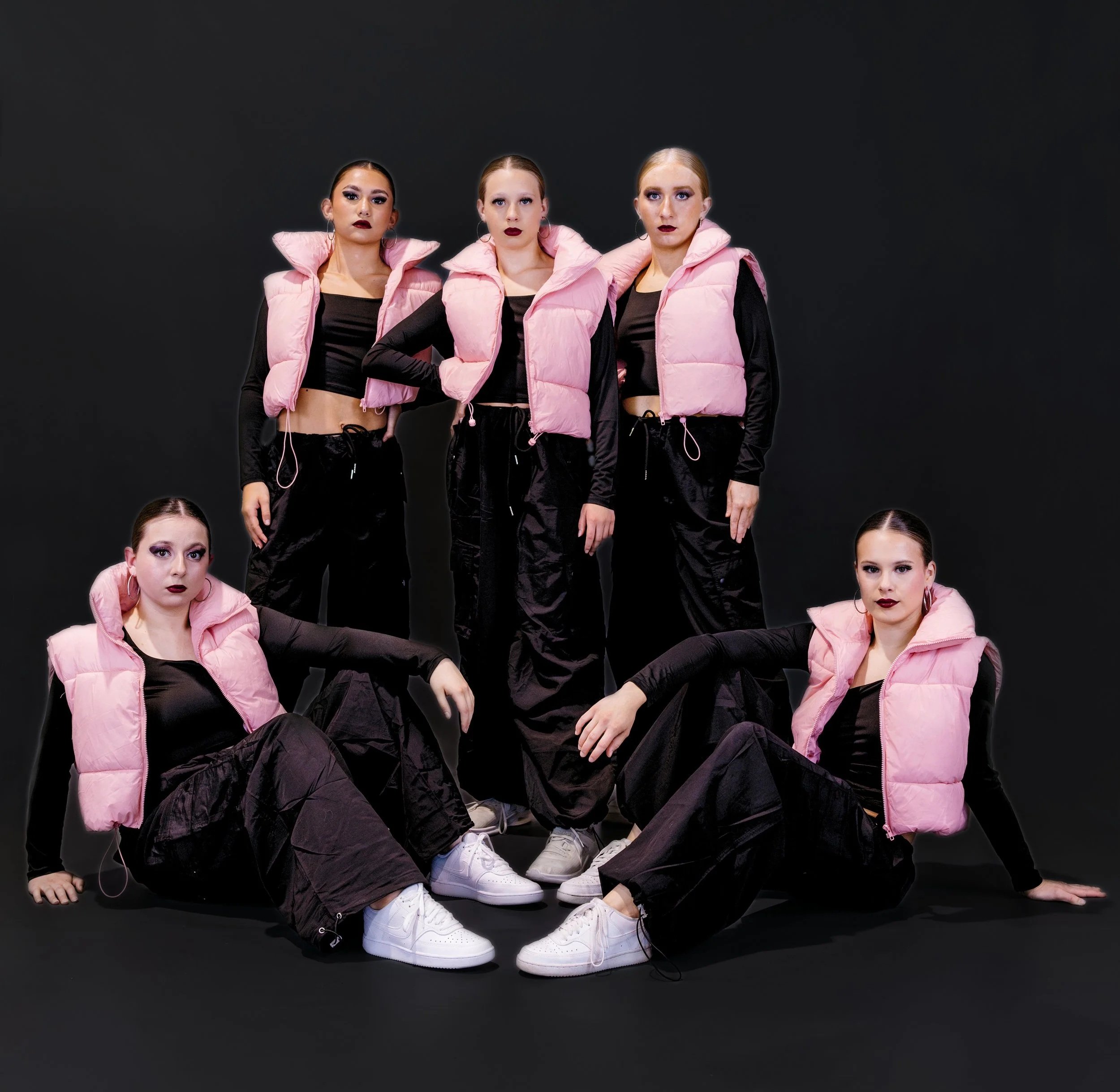 Three women standing in a row against a black background, wearing black athletic outfits with pink puffer vests, headbands, and hoop earrings, with serious facial expressions.