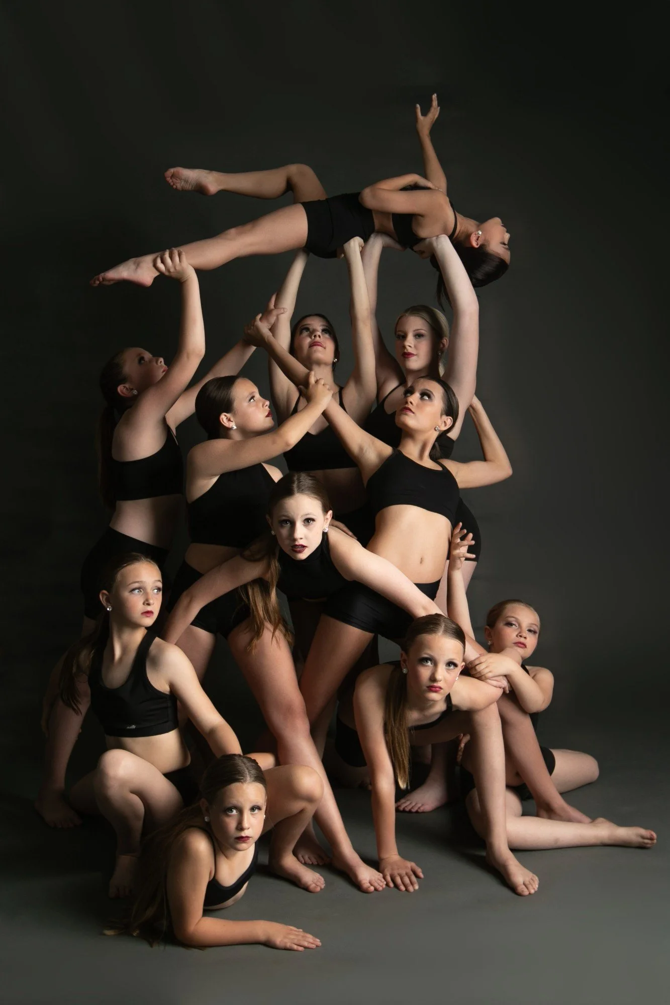 A group of young female dancers in black dancewear arranged in a human sculpture, with one dancer balanced horizontally on the backs of two others, against a dark background.