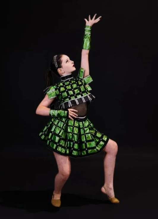 Young girl in a green and black geometric-patterned dance costume with matching arm accessories, posing with one arm raised and one hand on her waist, against a black background.