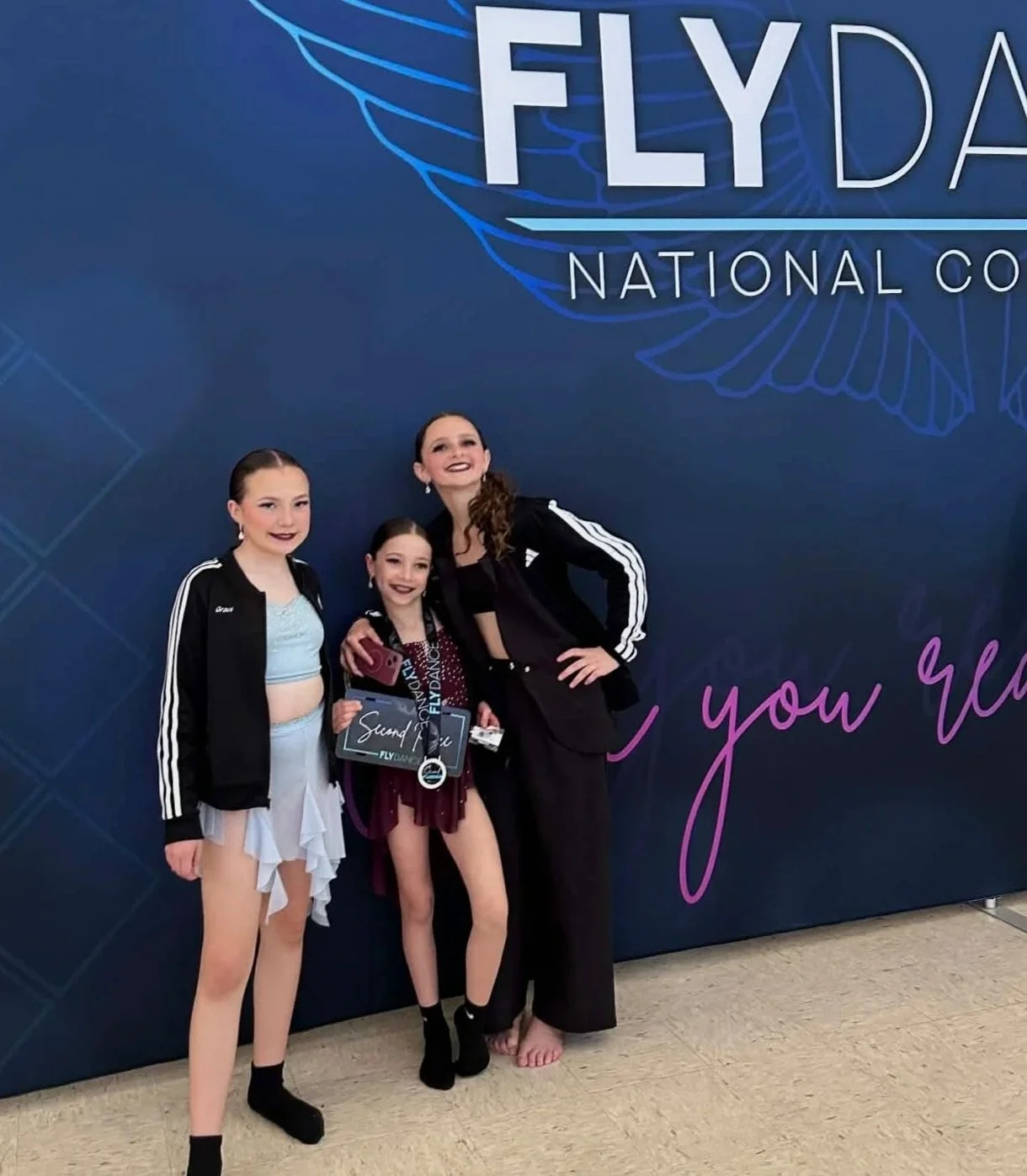 Three young female figure skaters and an adult woman standing together at a dance competition, posing in front of a blue backdrop with the words 'FLY DANCE' and 'NATIONAL COMPETITION' visible. The girl in the middle is holding a certificate and medals.