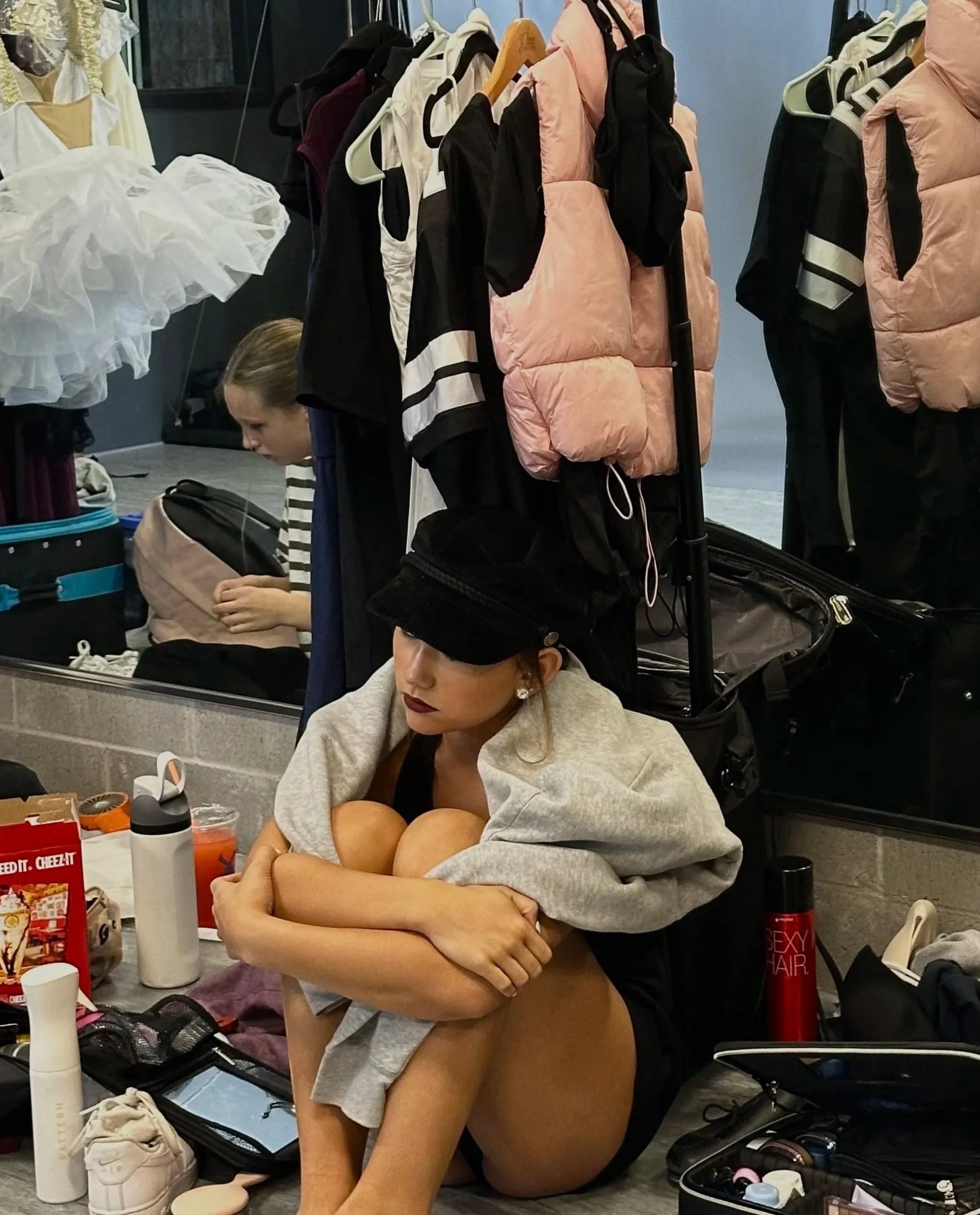 A woman is sitting on the floor with her arms wrapped around her knees, surrounded by makeup and hair styling products. She is partially undressed, wearing a gray hoodie and shorts, with makeup and earrings. In the background, there are clothing racks with jackets, dresses, and a person working on hair or makeup.