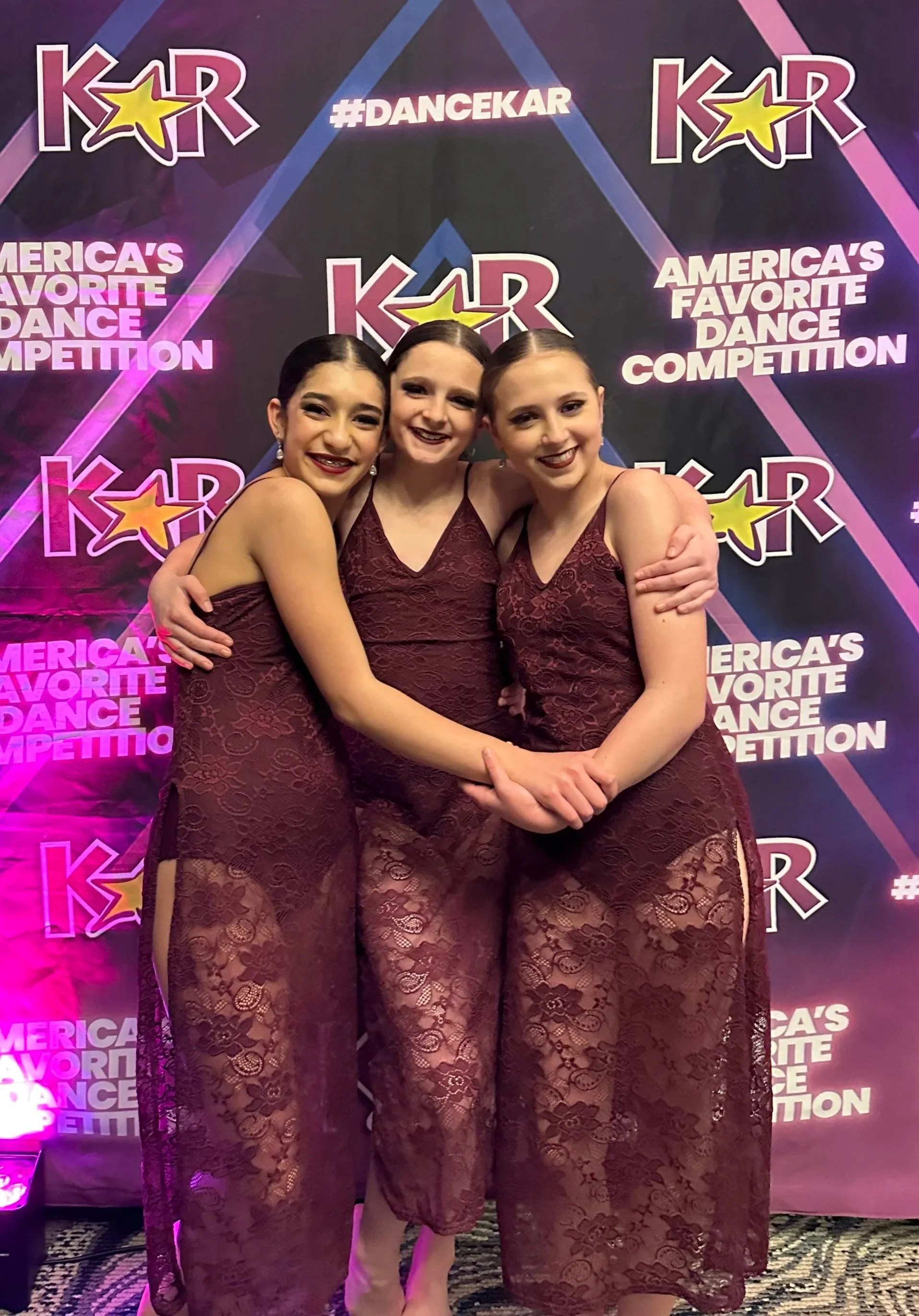 Three young female dancers in matching burgundy lace performance dresses standing in front of a backdrop with the text 'America's Favorite Dance Competition' and the hashtag '#DanceKar'. They are smiling and hugging each other.