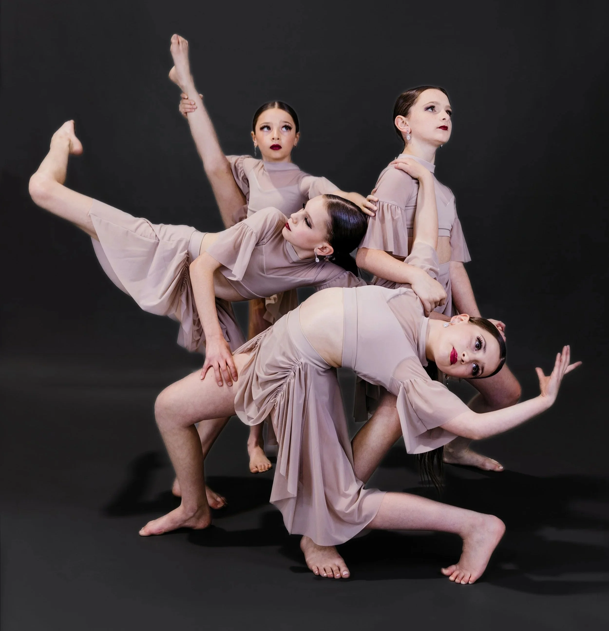 Four women in beige dresses performing an acrobatic pose against a black background. They are arranged with some lying on the ground and others balancing on their legs, creating a layered, artistic composition.