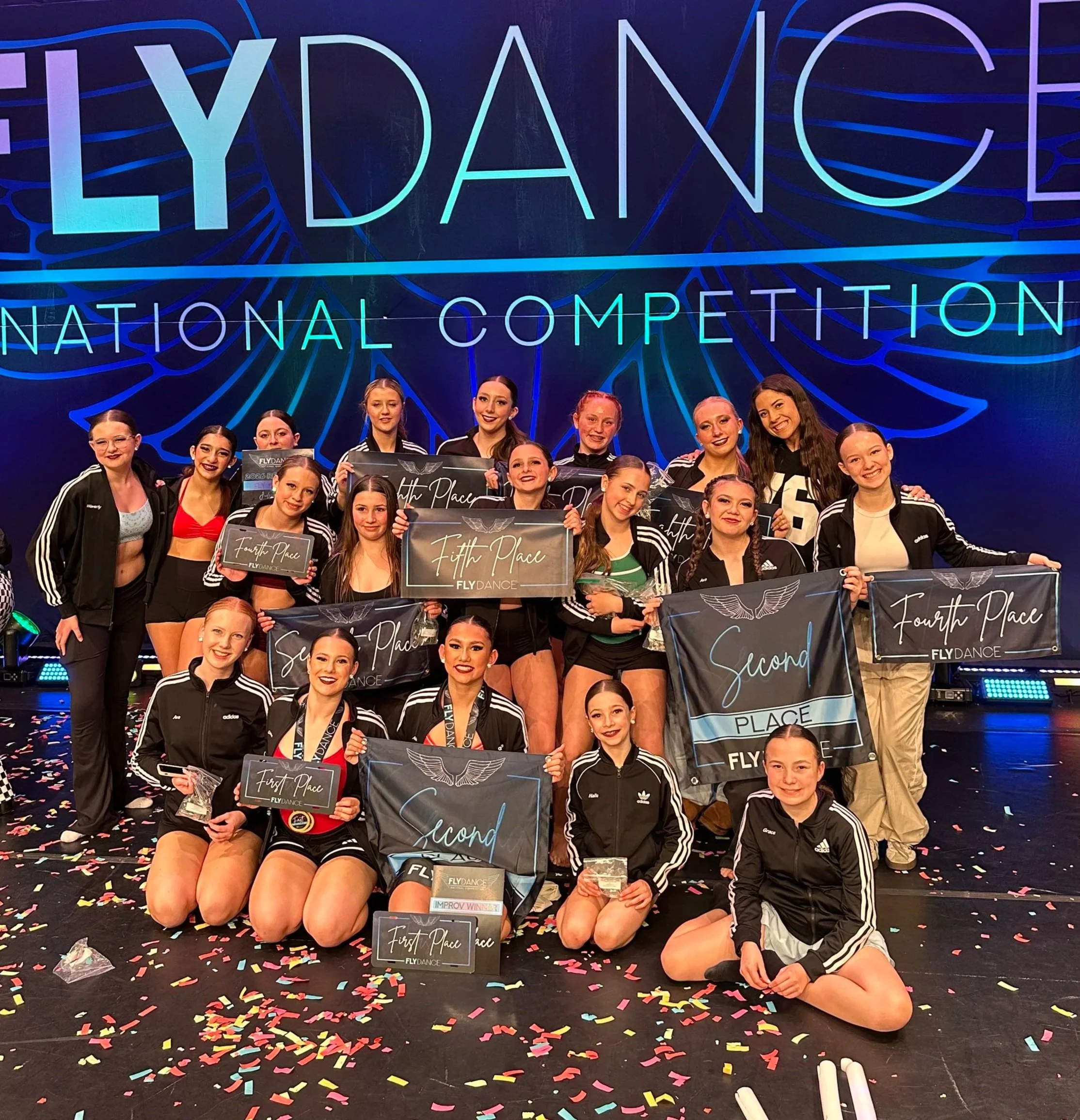Group of young female dancers in black athletic wear holding awards on stage with colorful neon sign reading 'Fly Dance National Competition' behind them, decorated with scattered confetti.