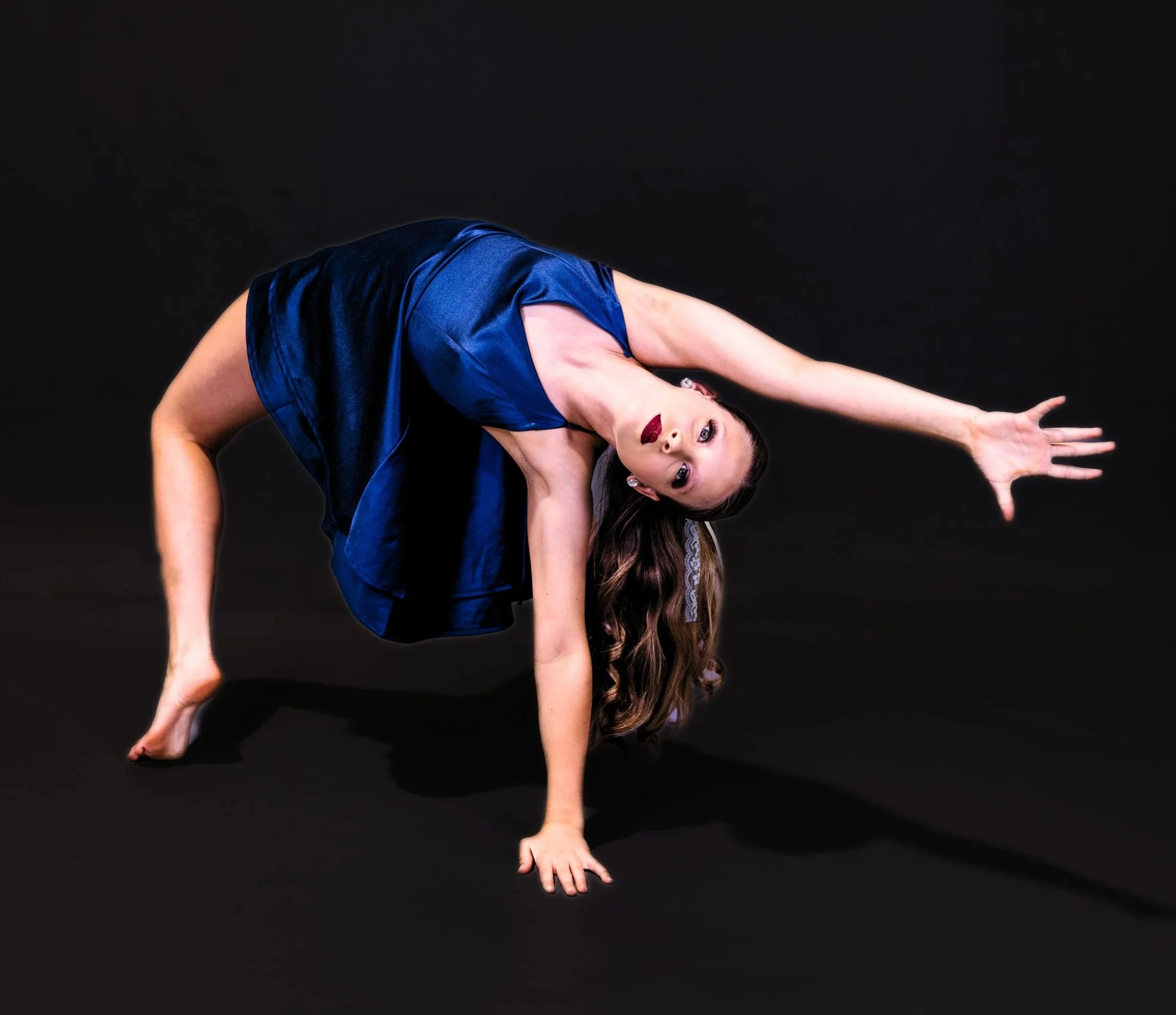 A woman with long brown hair in a dance pose on a black background, wearing a blue satin dress, with one hand on the ground and the other arm extended outward.