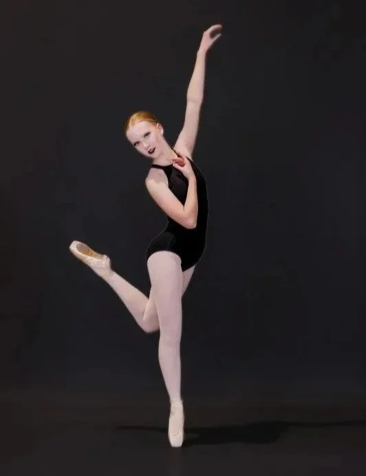 A young woman in a black leotard and white tights, performing a ballet pose against a black background.