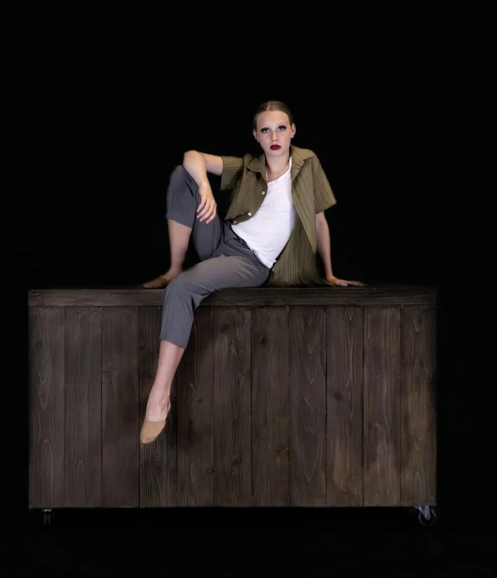 A woman with dark lipstick and eye makeup sitting on a wooden platform against a black background. She is wearing a green shirt, white t-shirt, gray pants, and beige high heels.