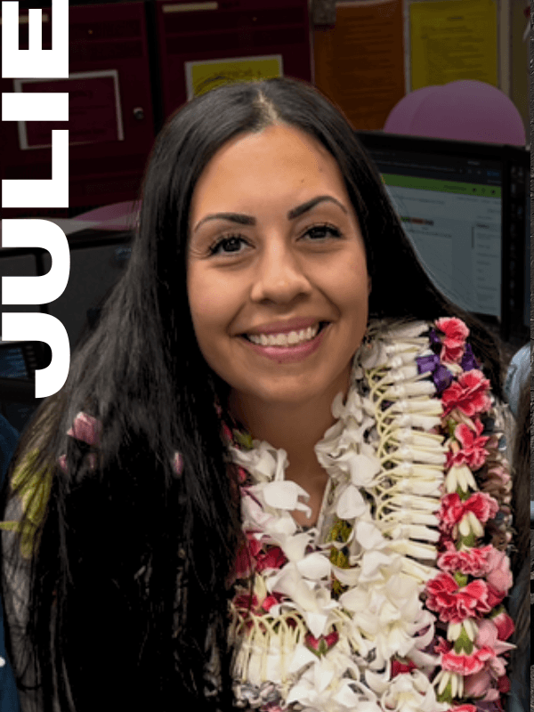 Young woman smiling, wearing a flower lei, sitting at a desk with a computer in the background, in an indoor setting.