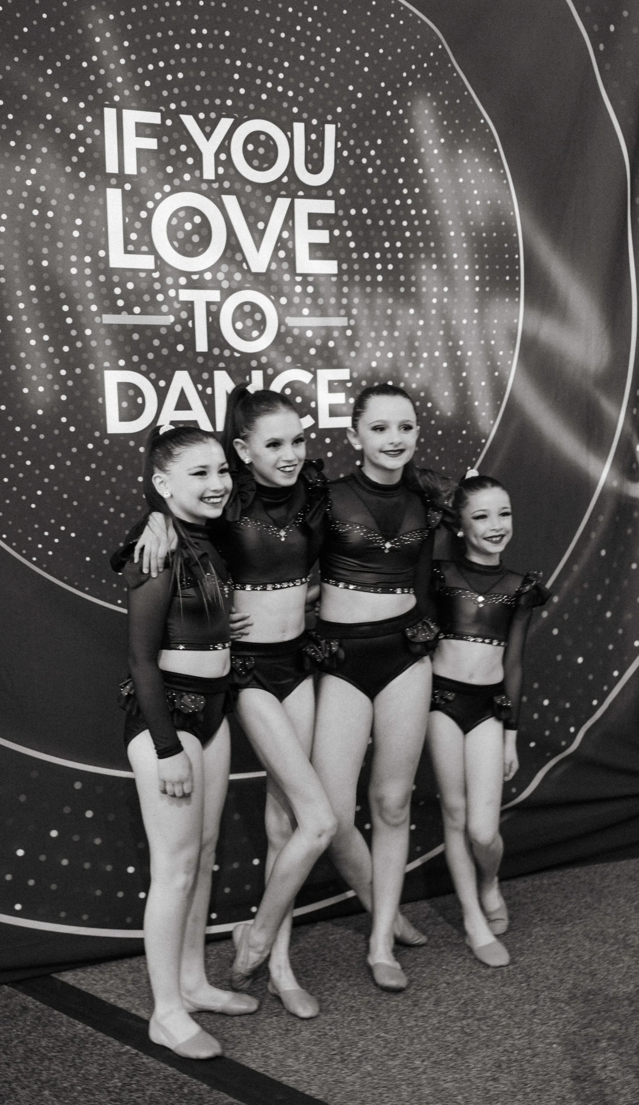 Four young female gymnasts in matching costumes standing together in front of a large sign that says "If you love to dance". They are smiling and posing with their arms around each other.
