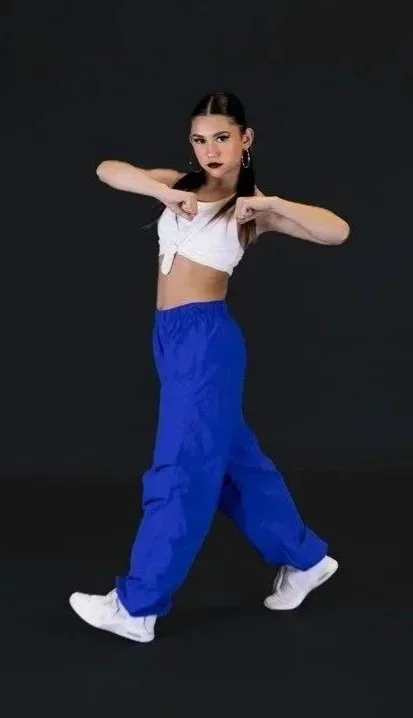 Young woman with dark hair, wearing a white crop top, blue baggy pants, and white sneakers, posing against a black background.