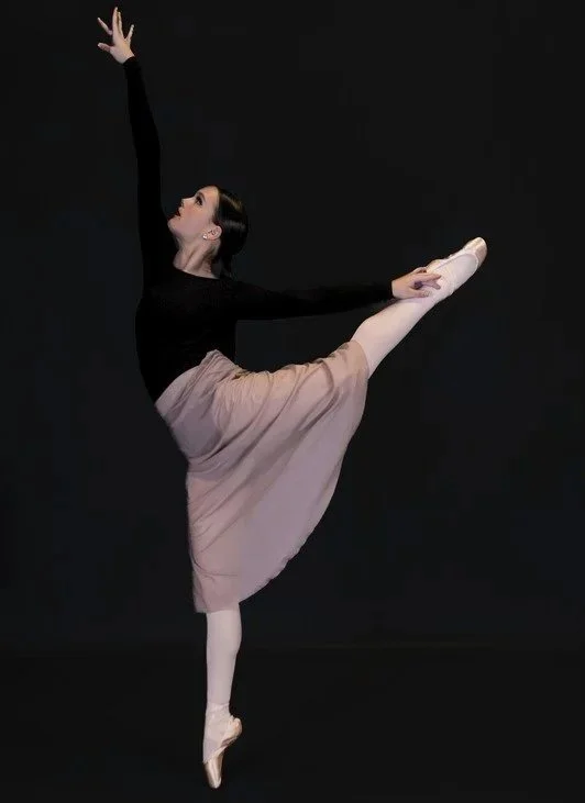 Ballet dancer in a black top and pink skirt performing a high leg extension against a black background.