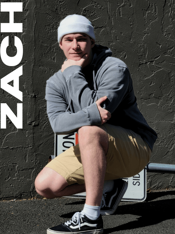 Young man wearing a white beanie, gray hoodie, khaki shorts, and black sneakers, sitting on a bench against a textured gray wall, with the word "ZACH" written vertically along the left side of the image.