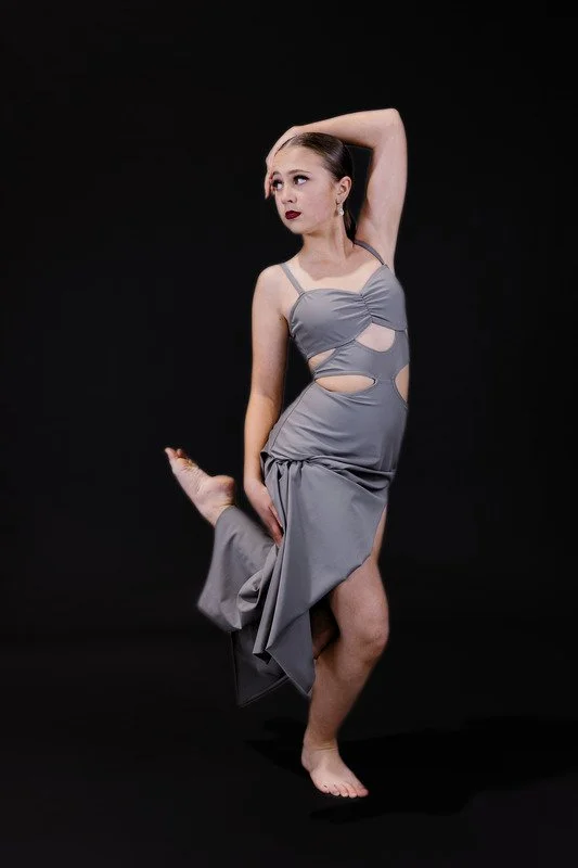 A young woman in a gray dress poses against a black background, with one arm behind her head and the other holding her leg, looking to the side.