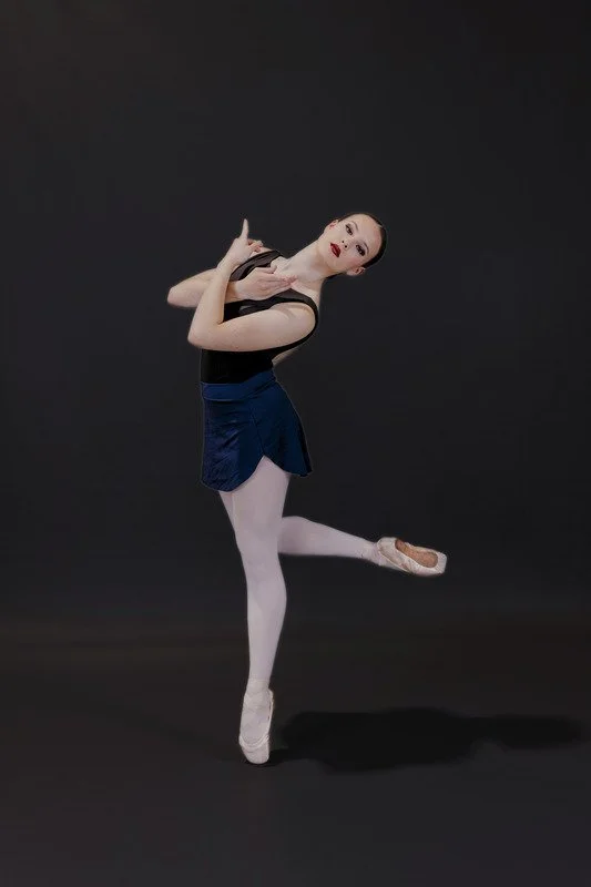 Ballet dancer in black leotard and blue skirt performing a pose against a dark background.