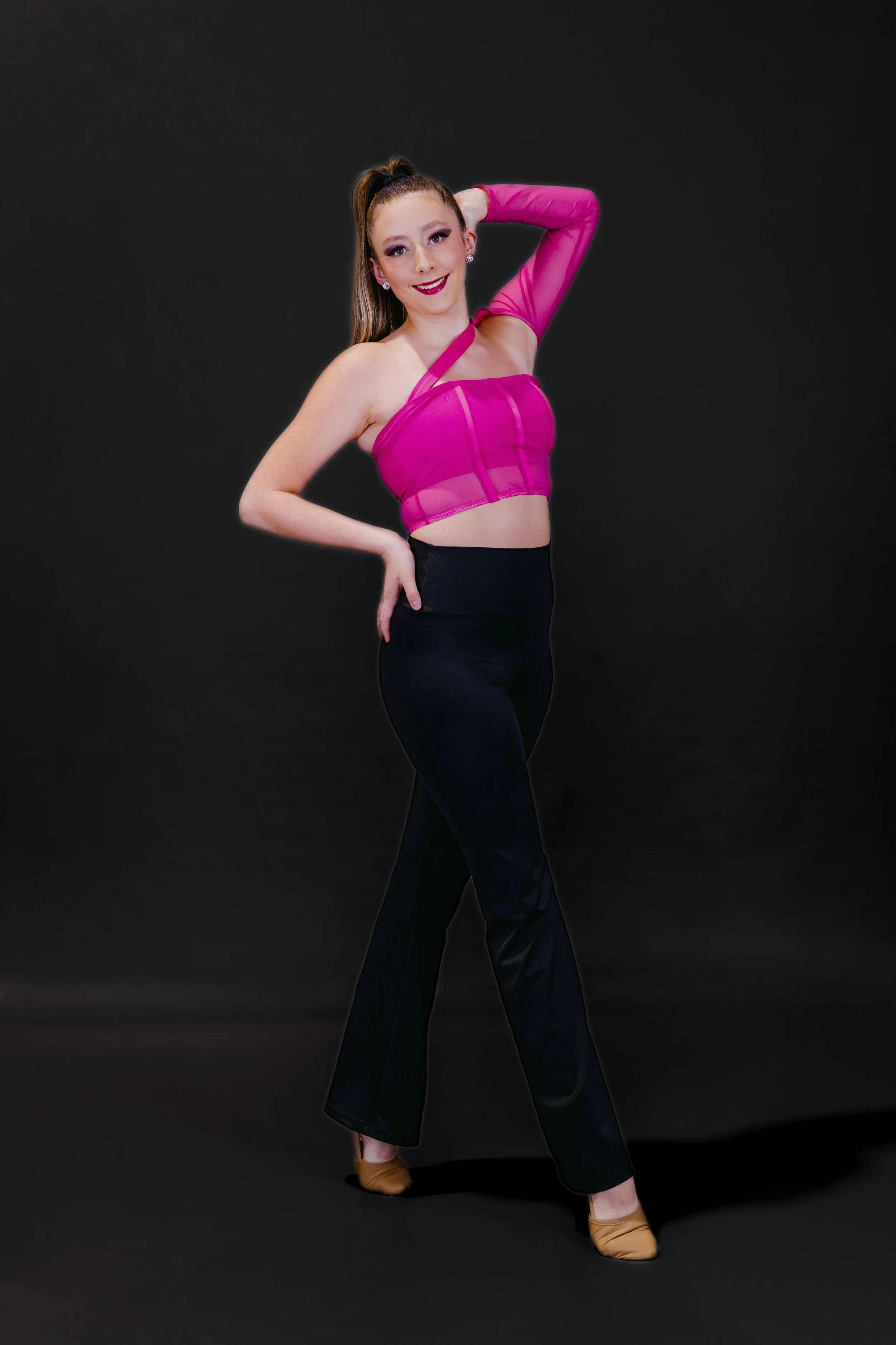 A woman in a pink sheer crop top, black pants, tan Latin dance shoes, posing with one hand on her hip and the other behind her head.